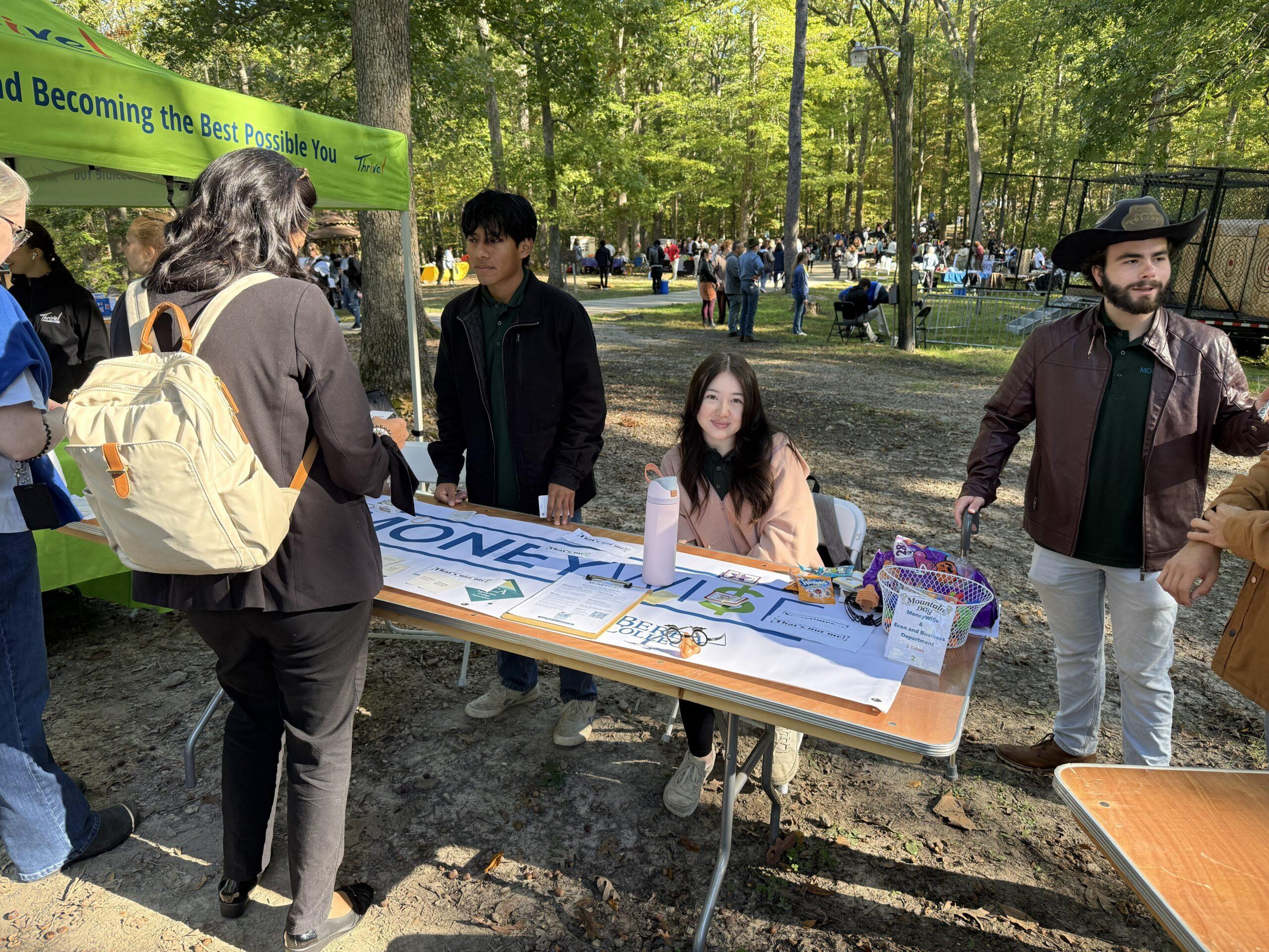 A female student sits at a table for the MoneyWi$e program and smiles for the camera and a male student stands beside her, speaking to interested peers across the table.