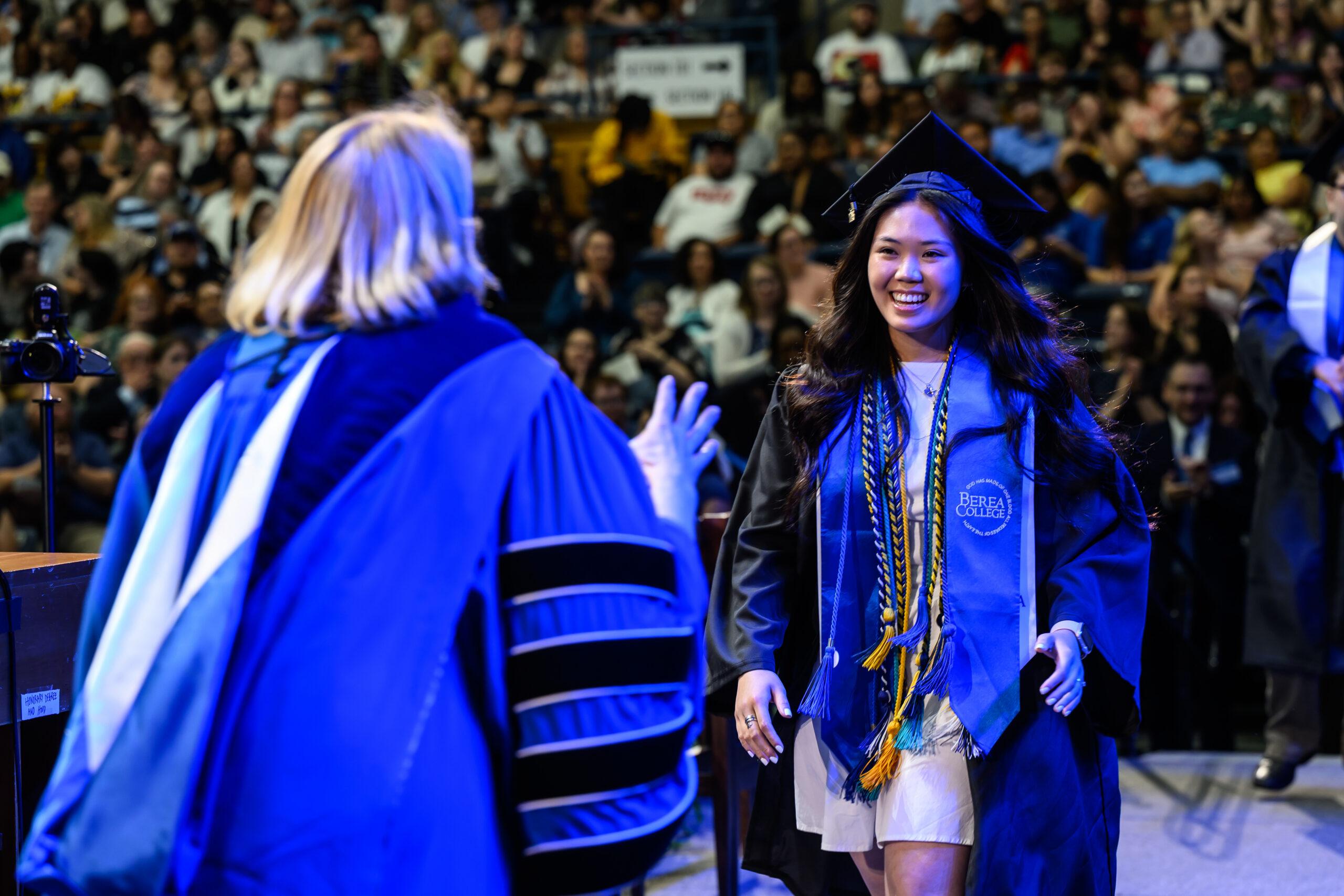 A female student dressed in graduation regalia smiles and walks toward Berea College president Cheryl Nixon onstage.