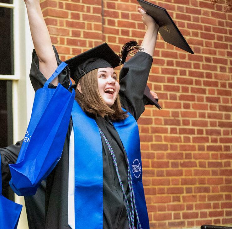 A student at graduation, in a cap and gown, throwing their hands in the air with joy.