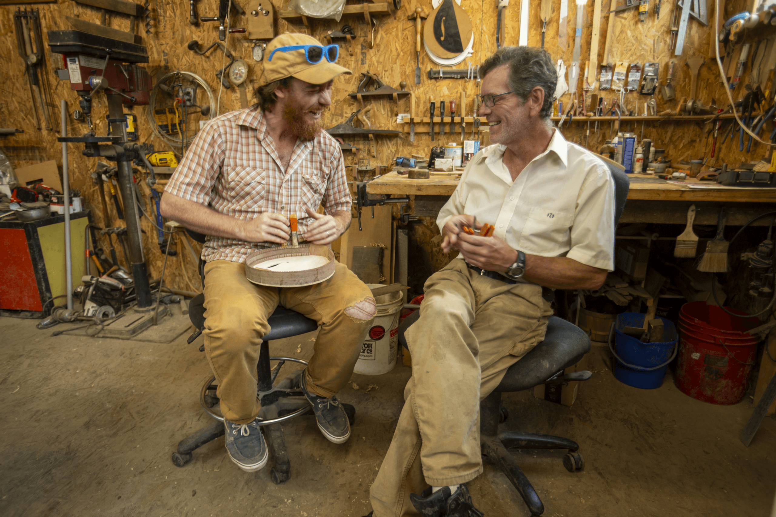 Two men sitting in a woodworking studio, laughing and working with their hands.