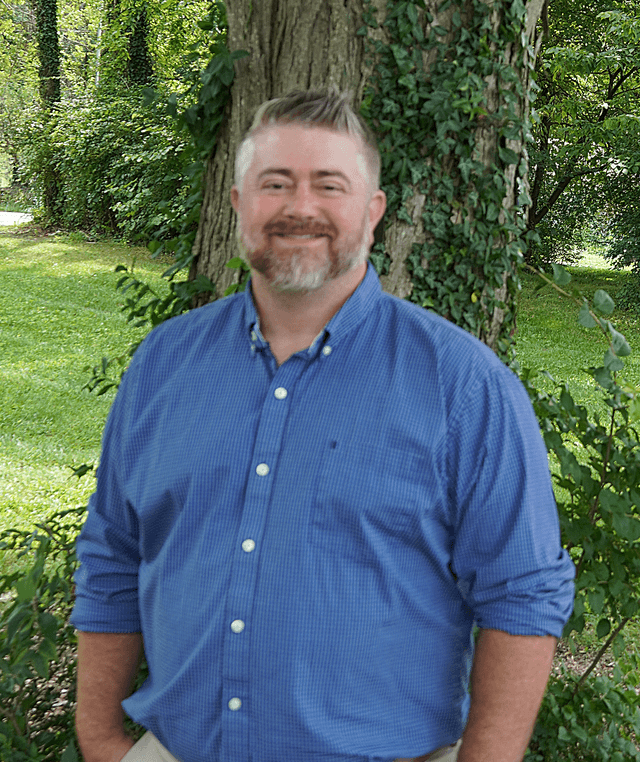 Brandon standing outdoors in front of a tree covered with ivy, wearing a blue button-down shirt.